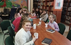 Grupa dzieci siedzi przy stole, dziewczynka spogląda przed siebie. W tle po prawej stronie regały z książkami. 