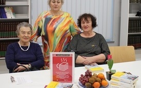 Dwie kobiety siedzą obok siebie, trzecia stoi nad nimi. Na stole owoce i plakat Dyskusyjnych Klub&oacute;w Książki. 