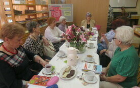 Grupa kobiet siedzi przy stole, na kt&oacute;rym są talerze, bukiet sztucznych kwiat&oacute;w. W tle logo Dyskusyjnych Klub&oacute;w Książki. 