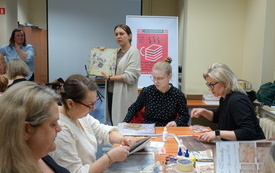 Kobiety siedzą przy stole i wycinają. W tle kobieta prowadząca warsztaty.