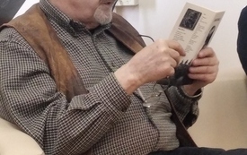 Starsszy mężczyzna w koszuli i kamizelce siedzi na fotelu trzymając otwartą książkę. 