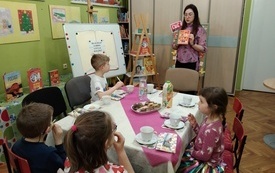 Bibliotekarka pokazuje książkę dzieciom, kt&oacute;re siedzą przy stole. 
