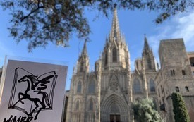 Dłoń trzymająca kartkę z logotypem Wojew&oacute;dzkiej i Miejskiej Bbilioteki Publicznej w Rzeszowie na tle katedry świętej Eulalii w Barcelonie