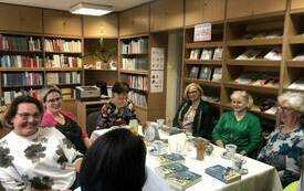 Grupa kobiet siedzi przy stole, na kt&oacute;rym są książki. W tle regały z książkami. 