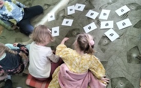 Na dywanie karteczki na kt&oacute;rych znajdują się ślady st&oacute;p. Obok grupa dzieci.