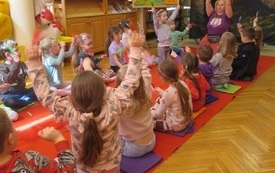 Grupa dzieci siedzi na czerwonym dywanie w bibliotece z uniesionymi do g&oacute;ry rękami w kt&oacute;rych trzyma pi&oacute;rka. 