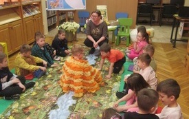 Chłopiec w ż&oacute;łto pomarańczowej pelerynie siedzi na środku dywanu, wok&oacute;ł grupa dzieci