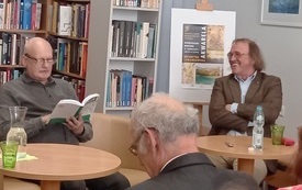 Przy stolikach dwóch starszych mężczyzn, jeden z mężczyzn trzyma w ręku książkę.