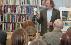 Starszy mężczyzna z książką w ręku stoi przed zebraną publicznością