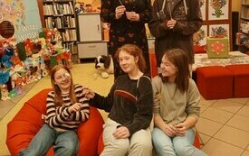Grupa nastolatk&oacute;w siedzi na pufach, za nimi młoda dziewczyna i bibliotekarka stoją. W tle regały z książkami. 