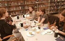 Grupa młodych ludzi siedzi przy stole, w tle regały z książkami. 