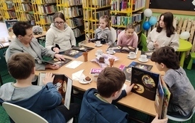 Grupa dzieci i bibliotekarka siedzą przy stole między regałami.