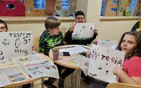 Grupa dzieci prezentuje prace plastyczne przy stolikach. 