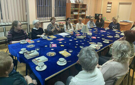 Grupa ludzi siedzi wokół stołu, który przykryty jest suknem, są na nim ksiażki i filiżanki.