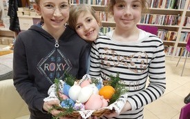 Trzy dziewczynki trzymają koszyk z jajkami. W tle regały z książkami. 