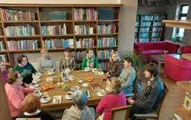 Grupa ludzi siedzi przy stole wok&oacute;ł regały z książkami.