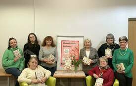 Grupa kobiet stoi blisko siebie, dwie siedzą i trzymają książki.