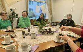 Grupa kobiet siedzi przy stole, na kt&oacute;rym są talerze, wazon z kwiatami. 