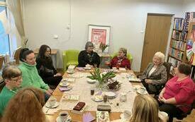 Grupa kobiet siedzi wok&oacute;ł stołu, w tle drzwi. 