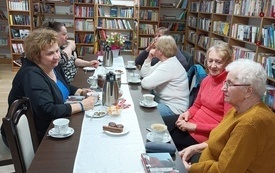 Gruppa kobiet siedzi przy stole, wok&oacute;ł regały z książkami.