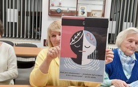 Trzy kobiety siedzą obok siebie, kobieta w środku trzyma kartę z gry "Świteź".