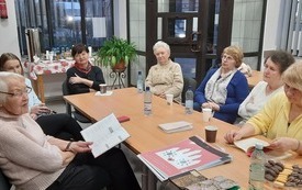 Grupa kobiet siedzi przy stole, jedna z nich trzyma książkę na kolanach.