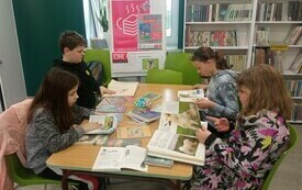 Czworo dzieci siedzi i przegląda książki z ilustracjami, w tle regał z książkami.