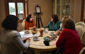 Kilka kobiet siedzi przy stole i rozmawia, w tle okno. 