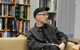 Mężczyzna pisarz w czapce z daszkiem, okularach i kolczykiem w uchu. W tle książki. 