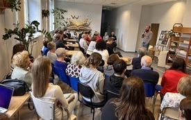 Grupa ludzi siedzi na krzesłach, przed nimi pisarz Wojciech Chmielarz trzyma mikrofon i mówi.