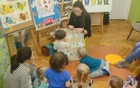 Bibliotekarka pokazuje planszę, dzieci podchodzą i wskazują na ilustracje.