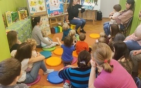 Bibliotekarka czyta książkę i pokazuje ilustracje, wokół dorośli i dzieci.