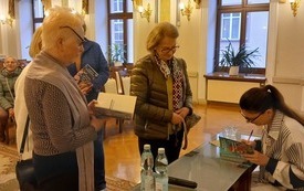 Grupa kobiet stoi z książkami obok Ałbeny Grabowskiej, autorka wpisuje dedykacje dla czytelniczki. 