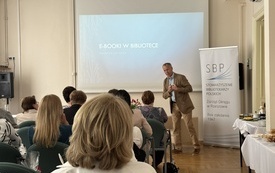 Grupa osób w różnym wieku siedzi i ogląda na ekranie prezentację multimedialną przedstawianą przez mężczyznę w garniturze