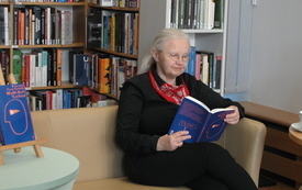 Starsza kobieta czyta książkę siedząc na kanapie obok p&oacute;łek z książkami