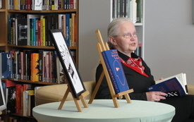 Starsza kobieta siedzi na kanapie obok p&oacute;łek z książkami, w ręce trzyma książkę