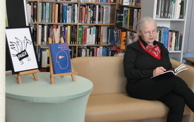 Starsza kobieta siedzi na kanapie obok p&oacute;łek z książkami, na kolanach trzyma otwartą książkę