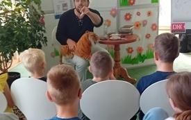 Mężczyzna mówi do dzieci siedzących przed nim w tle baloniki.