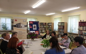 Grupa kobiet rozmawia przy stole, w tle plakat i logotypy Dyskusyjnych Klub&oacute;w Książki.
