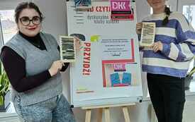 Dwie nastolatki trzymają książki i stoją przy planszy z logo DKK. 