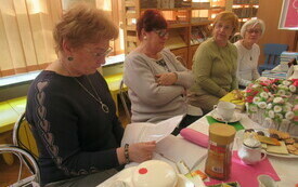 Cztery kobiety siedzą przy stole, jedna z nich trzyma zadrukowaną kartkę papieru i pochylona czyta. 