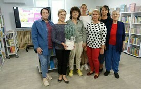 Siedem kobiet stoi obok siebie, jedna z nich trzyma doniczkę z rośliną. W tle regały z książkami.