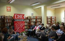 Na fotelu siedzi Jakub Małecki, pisarz, na drugim fotelu Dyrektor biblioteki  w Lubaczowie, rozmawiają, w tle grupa ludzi, siedzi na krzesłach. 