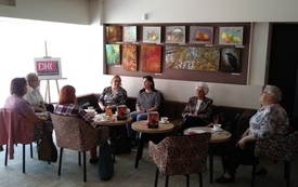 Grupa ludz siedzi dookoła stołu, w tle obrazy na ścianach i logo DKK. 