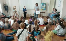 Kobieta i mężczyzna stoją, reszta siedzi i słucha.