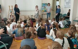 Kilkanaście dzieci siedzi na dywanie, mężczyzna na przeciwko z kobietami, jedna z nich mówi do zgromadzonej publiczności.