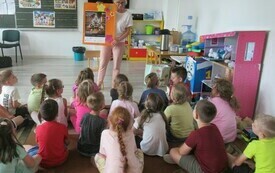 Grupa dzieci siedzi na podłodze w klasie szkolnej patrząc na bibliotekarkę prezentującą plakat Og&oacute;lnopolskiego Tygodnia Czytania Dzieciom