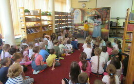 Grupa dzieci siedzi na podłodze w bibliotece, przed nimi stoi aktorka Teatru Kokon