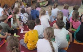 Grupa dzieci siedzi na podłodze w bibliotece słuchając czytanej przez aktora bajki