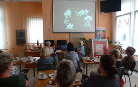 Grupa kobiet siedzi przy stolikach w bibliotece i ogląda prezentację multimedialną na ekranie